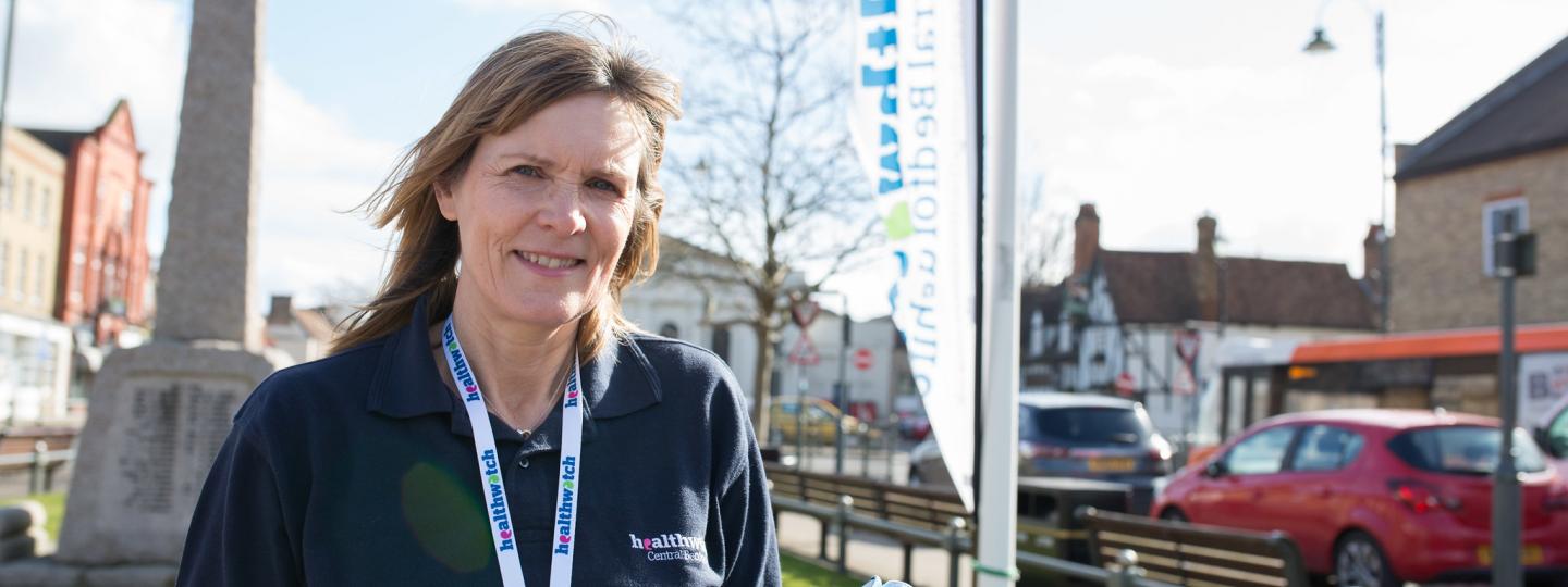 Female volunteer wearing a Healthwatch t shirt and lanyard standing in front of a flag